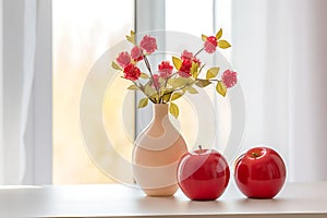 On a daytime day there is a vase with red flowers and artificial apples on the windowsill