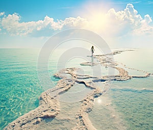Silhouette di una giovane donna che cammina su di sali del Mar Morto riva al sorgere del sole verso il sole.