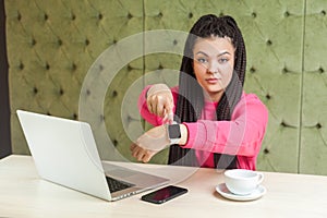 Deadline! Portrait of serious young bossy girl with black dreadlocks hairstyle in pink blouse sitting showing time on her