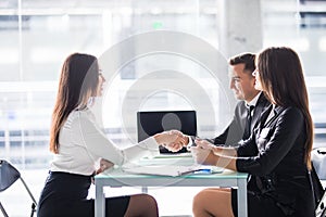 Deal. Side view of cheerful young man sitting close to his wife and shaking hand to woman sitting in front of him at the desk offi