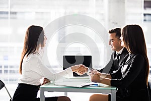 Deal. Side view of cheerful young man sitting close to his wife and shaking hand to woman sitting in front of him at the desk offi