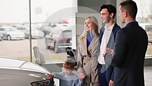 dealership, happy young male and female buyers with a child choose a family car in a showroom and consult with a manager