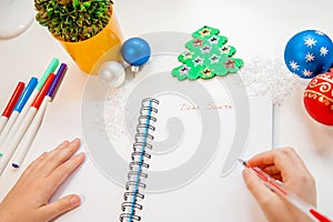 Dear Santa letter, Christmas card. A child holding a pen writes on a white sheet on a wooden background with New Year`s decor