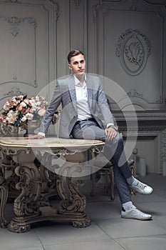 Dear young man with a haircut in a suit, sitting at a table in a room with a classic interior. Luxury. Male beauty