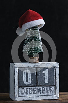 December 01 on wooden calendar standing next to Christmas tree decorated with santa claus hat.