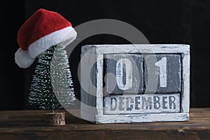 December 01 on wooden calendar standing next to Christmas tree decorated with santa claus hat