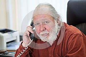 Decisive senior manager holding telephone handset and calling in office room, white beard and grey hair, looking at camera