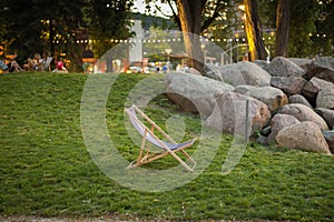 Deck chair standing on green grass at sunset in Garnizon with rocks, trees and blurred people in the background