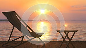 Deck chair and table with glass of wine at lake coast against orange sunset