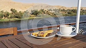 The deck of a cruise ship. On a wooden table there is a cup of coffee and a saucer with pastries