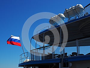 Deck of a pleasure yacht or ship, with seats, fencing and equipment. The Russian flag flutters in the wind. Blue sky