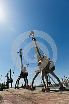 Decommissioned port cranes tower over abandoned dockyard under blue sky. Maritime cargo loaders, signs of urban decay in