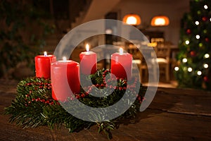 Decorated Advent wreath from fir branches with red burning candles on a dark wooden table, living room with Christmas tree blurred