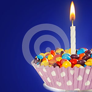 Decorated Birthday cupcake with one lit candle and colorful candies on blue background