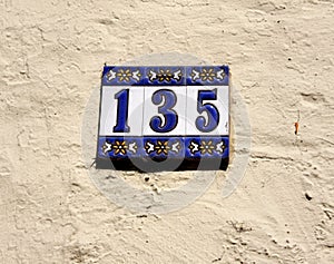 Decorated blue and white house address number 135, hung on a bright, rough wall