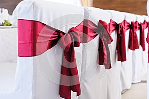 Decorated chairs made of white fabric and large burgundy satin bows. White fabric chair covers stand in a row