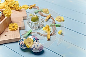 decorated Easter eggs, yellow flowers, burning candle and brown boards on wooden blue background. Spring and Easter concept