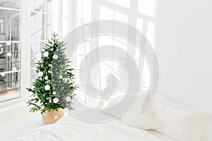 Decorated festive Christmas tree in white classic bedroom interior with soft bed. Winter home interior. Cozy season. New Year,