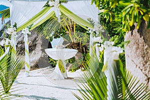 Decorated romantic wedding celebration location, table and chairs on tropical beach. Lush green foliage and white lowers