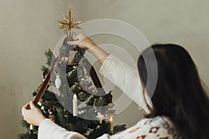 Decorating christmas tree with stylish golden star and ribbon. Woman in cozy sweater hanging vintage ornament on tree branch close