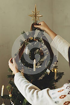 Decorating christmas tree with stylish golden star and ribbon. Woman in cozy sweater hanging vintage ornament on tree branch close