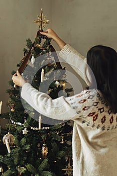 Decorating christmas tree with stylish golden star and ribbon. Woman in cozy sweater hanging vintage ornament on tree branch close