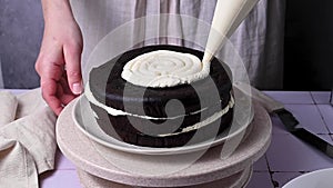 Decorating home baked chocolate cake. Female hands piping out whipped cream on a sponge cake.