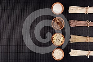 Decorative border of bundles raw noodles with ingredient in wooden bowls on black striped mat background with copy space, top view