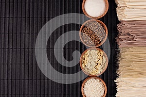 Decorative border of bundles raw noodles with ingredient in wooden bowls on black striped mat background with copy space, top view