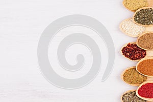 Decorative border of different spices in bamboo spoons on white wood background, top view, closeup, vertical.