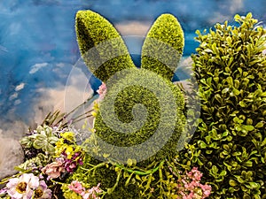Decorative green topiary rabbit head with flowers around its neck and surrounded by greenery in front of blue sky-like background