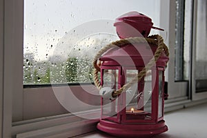 Decorative lantern pink with a burning candle inside, at home on the window. rainy autumn day and rain drops on PVC
