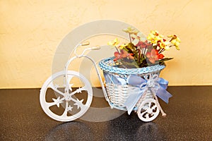 Decorative plastic tricycle with a beautiful basket, bow and flowers standing on a brown shelf. Interior design