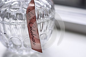 Decorative retro glass jar container with a pattern with a pink ribbon with text Lets Celebrate on a white windowsill
