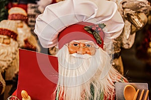 Decorative Santa doll in chef hat holding blank sign and cooking implements - close-up and selectie focus - other blurred santas