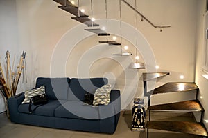 Decorative stairs in the apartment made of bronze material. Above them is a light from lamps. A sofa under the stairs. The concept