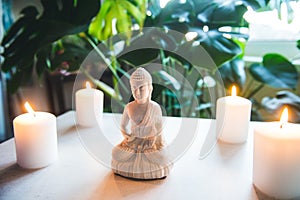 Decorative white Buddha statuette with candles and green monstera plant on the background. Meditation and relaxation