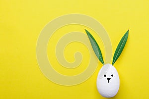 Decorative White Painted Easter Egg Bunny with Drawn Cute Kawaii Face. Green Leaves as Ears. Pastel Yellow Background. Spring