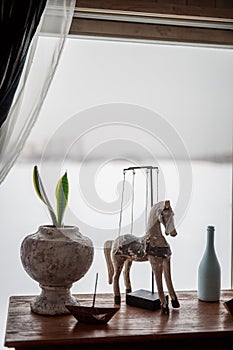 Decorative wooden horse stands on the table near the window flowers in pots, white bottle, iron boat, elements of room decoration,