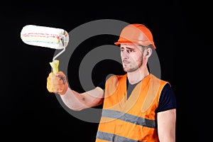 Decorator painting surface in front of himself. Man in helmet, hard hat holds paint roller, black background. Paint and