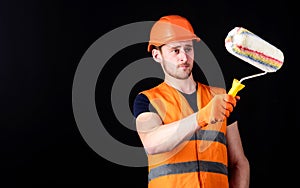 Decorator painting wall, copy space. Man in helmet, hard hat holds paint roller, black background. Worker, painter