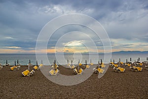 Deep Cloudy Blue Sunset Sky with Yellow Lights above Water and Many Folded Chaise-Lounges Umbrellas Beach