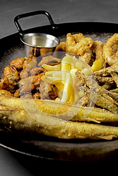 Deep fried set made from french fries, onion rings, big and small fried fish served in a wok over black background.