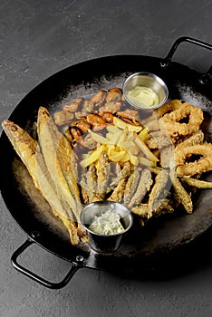 Deep fried set made from french fries, onion rings, big and small fried fish served in a wok over black background.