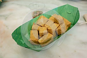 Deep fried tofu is cut into square balls in a green paper dish. The side has a cup of sauce.