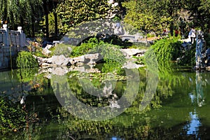 A deep green lake in a Japanese garden surrounded by lush green trees reflecting off the lake water