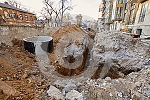 Deep and long trench dug out in ground near a modern new building under construction. Street with old houses and high