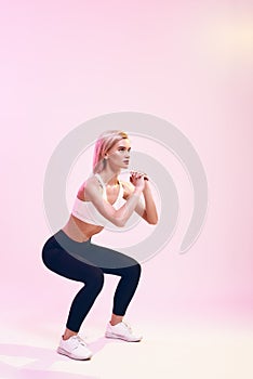 Deep squat. Back view of sporty and cute slim woman in sportswear doing squat while standing against pink background in