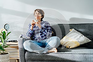 Deep in thoughts, dreaming woman drinking coffee sitting on sofa in sunlight beams after reading a book at home in cozy