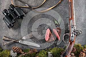 Deer or venison steak with antique long gun, cutlery, binocular and ingredients like sea salt, herbs and pepper, food background f